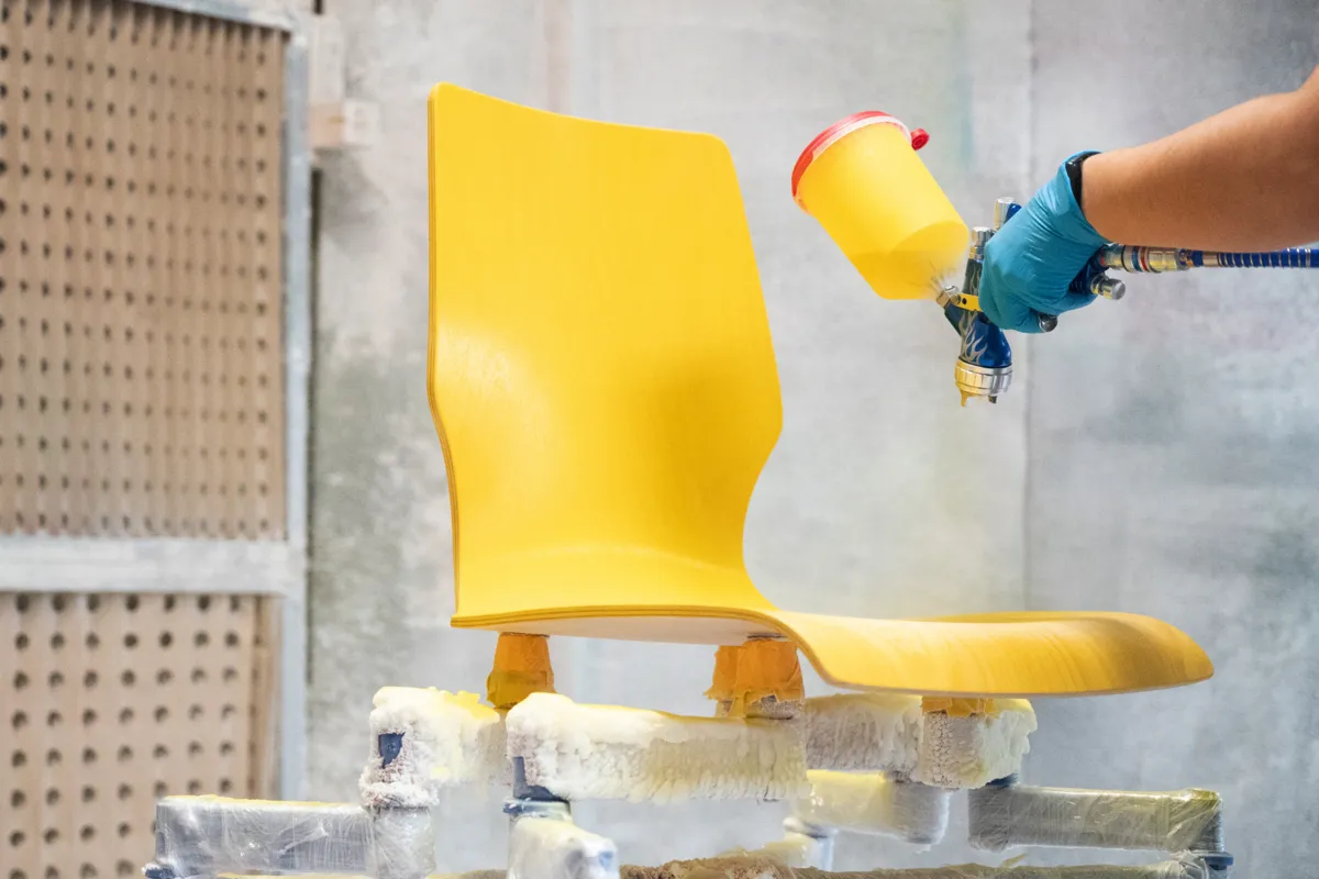 Lackierkabine bei ADUKA in Rothrist. Die ergonomische Sitz- und Rückenlehne wird mit einer Spritzpistole gelb lackiert. Der Stuhl ist auf einer Halterung fixiert.